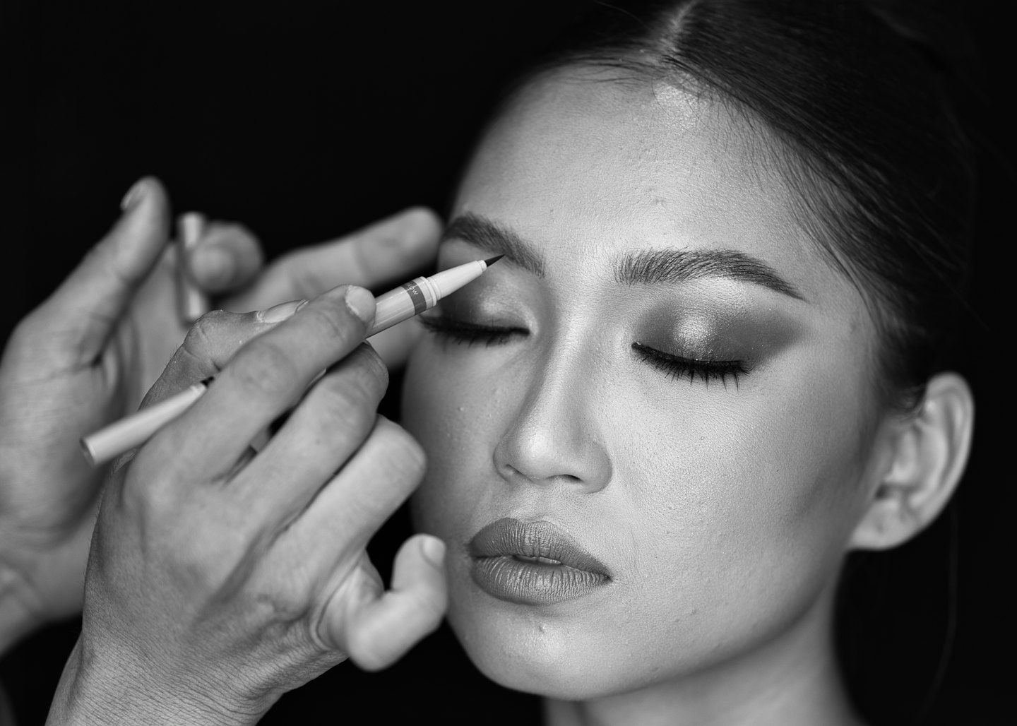 Makeup artist applies brow pencil to woman's closed eye.